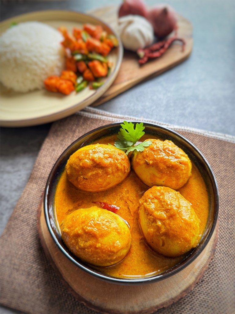 egg masala in a serving bowl with rice and a veggie at the side
