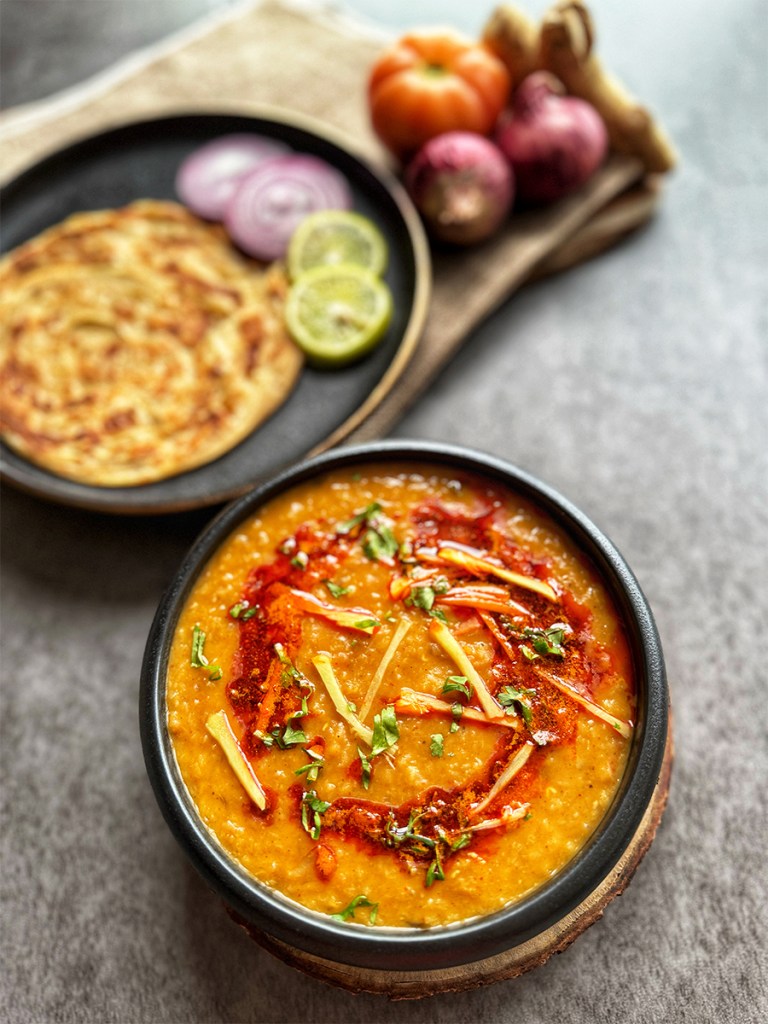 masoor dal tadka on a serving bowl with a serving of paratha, onions slices and lemon slices on the side
